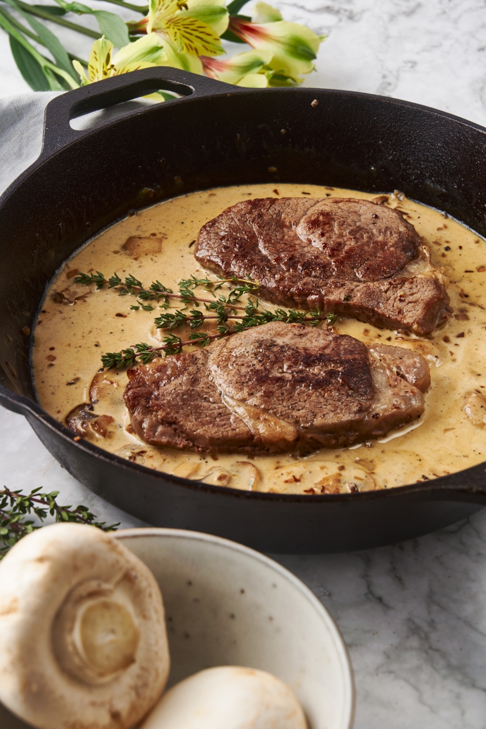 Two seared filet mignon steaks in a cast iron skillet surrounded by a brown mushroom sauce. There is a bowl of whole raw mushrooms next to the skillet.