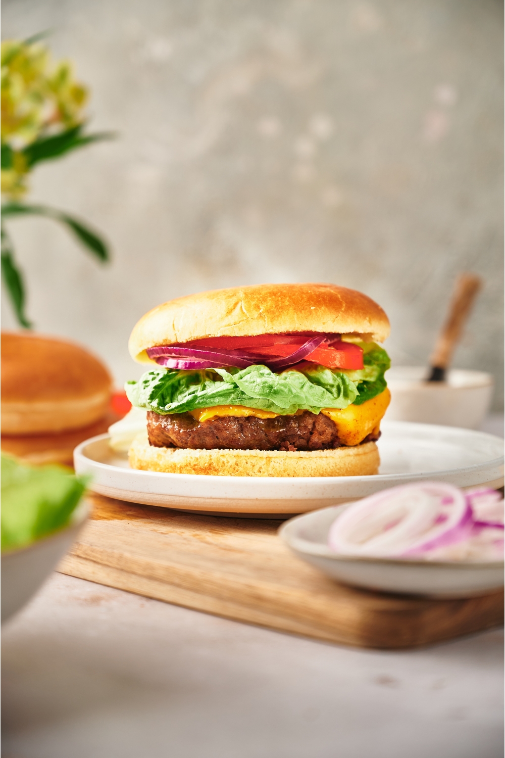 A cheeseburger with red onion, lettuce, and tomato on it. The burger is on a white plate atop a wood cutting board alongside a plate of sliced onions.