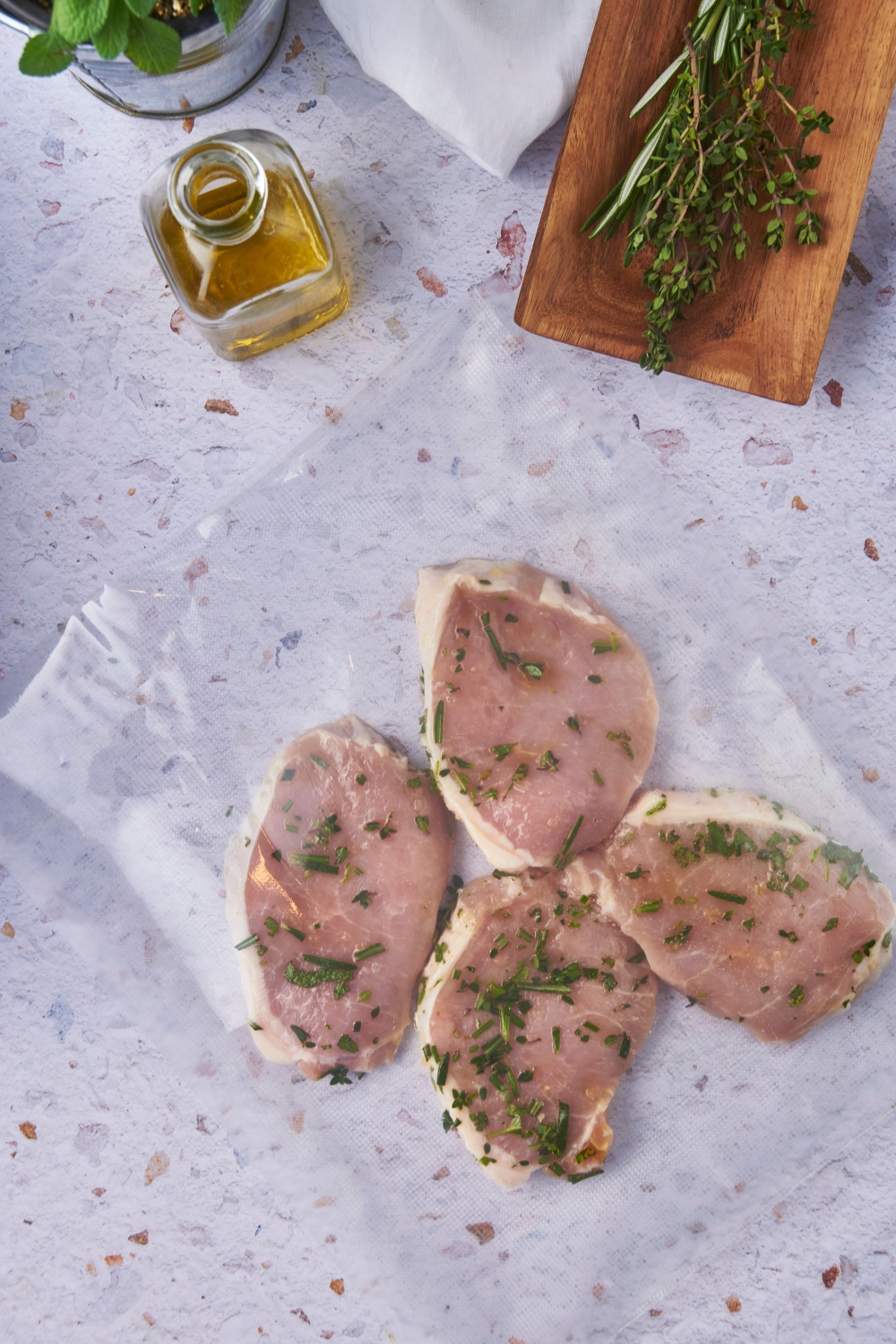 Four pork chops in a vacuum-sealed bag with fresh herbs. Next to the bag of pork chops is a container of oil and a bunch of fresh herbs.