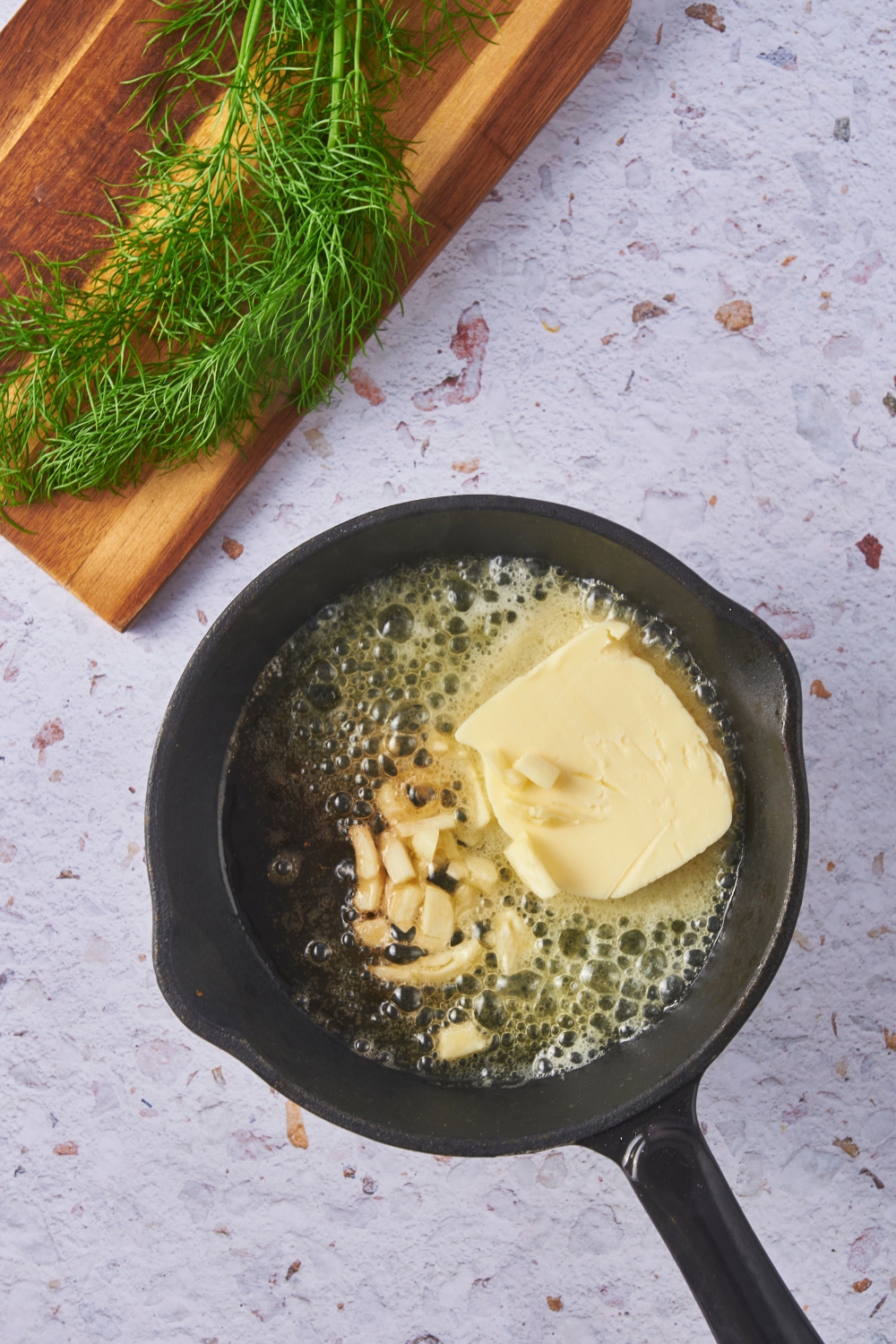 A black cast iron skillet with a large tab of melting butter and sliced garlic cloves in the skillet.