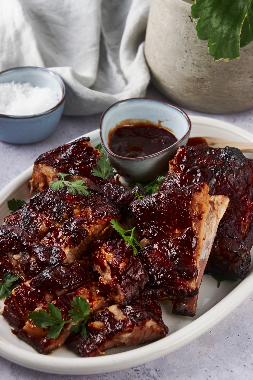 Cooked ribs covered in barbecue sauce piled on top of each other on a white plate, with a side of barbecue sauce on the plate and fresh parsley garnished on top of the ribs.