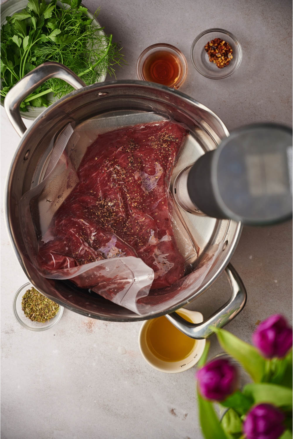 A tri-tip steak being sous vide in a water bath with a sous vide machine in the water bath. Surrounding the pot of water are bowls of seasonings and oil.