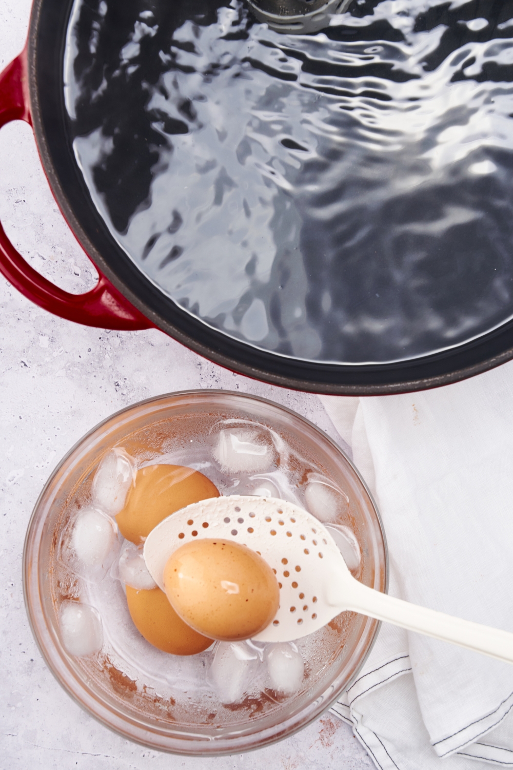 A slotted spoon lifting an egg out of an ice bath, next to a large pot of water.