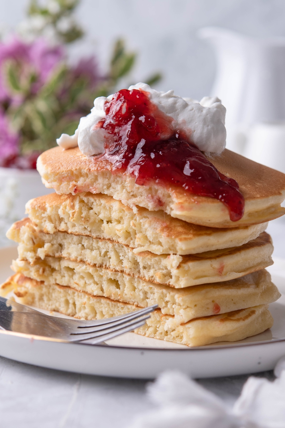A stack of 5 flapjacks on a plate topped with strawberry jam and a dollop of whipped cream. The stack is sliced almost in half to show the fluffy interior of the flapjacks.