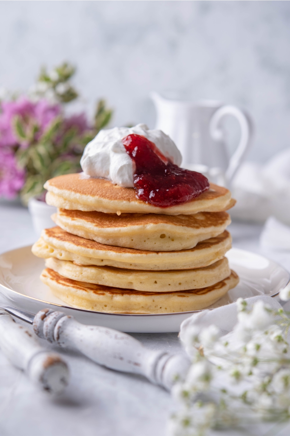 A close up of a stack of flapjacks topped with strawberry jam and whipped cream on a plate.