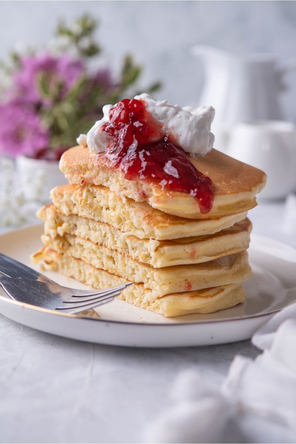 A stack of 5 flapjacks on a plate topped with strawberry jam and a dollop of whipped cream. The stack is sliced almost in half to show the fluffy interior of the flapjacks.