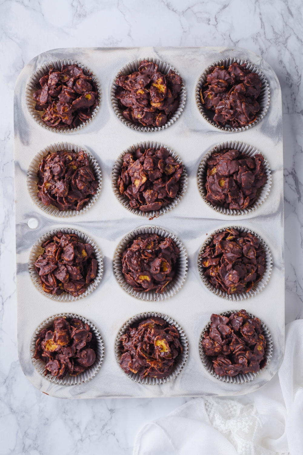 An overhead view of a muffin tin with chocolate cornflake clusters in muffin liners.