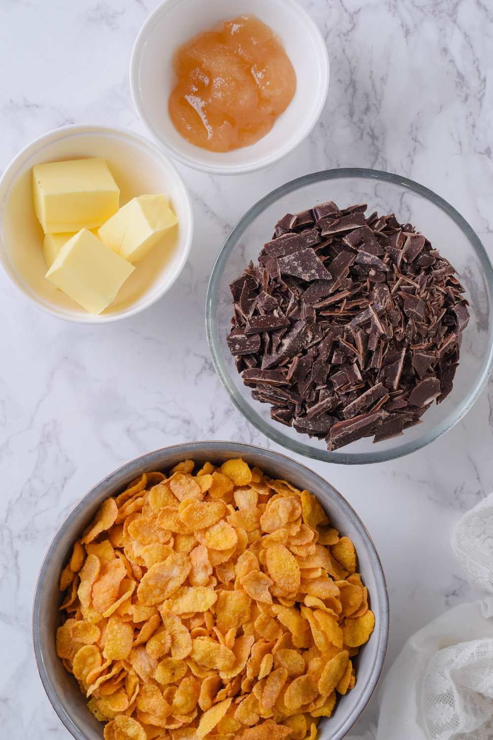 An overhead view of a bowl with cornflakes, chocolate chunks, butter, and honey.