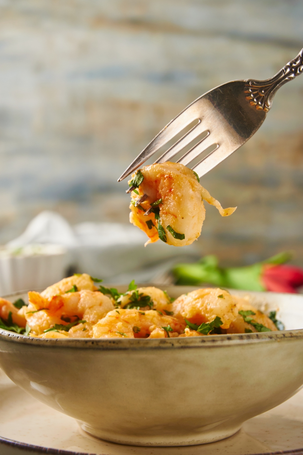 A fork holding a piece of cooked shrimp with fresh green herbs on it. The rest of the shrimp is in a bowl in the background.