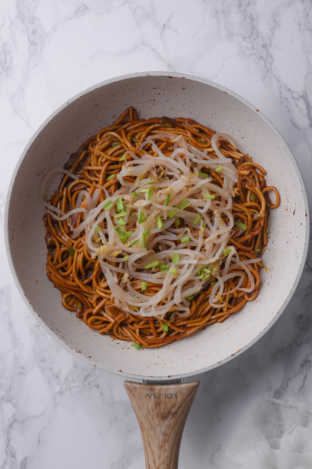 An overhead view of a skillet with pan fried noodles topped with bean sprouts and green onion slices.