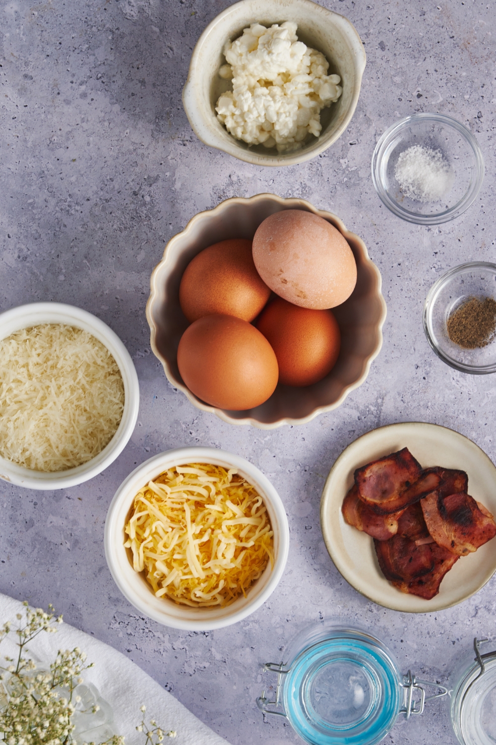 An overhead view of a bowl with whole eggs, a small bowl with cooked bacon, and three small bowls with various cheeses.