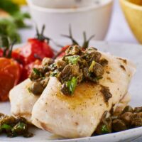 Two halibut fillets on a plate with capers on top of them.