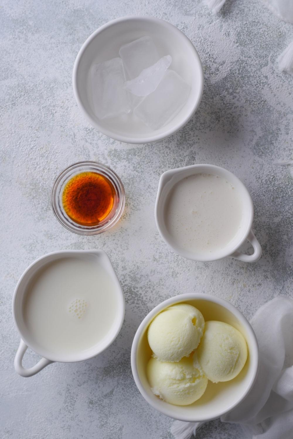 An overhead view of various bowls with ice cubes, vanilla extract, whole milk, and scoops of ice cream.