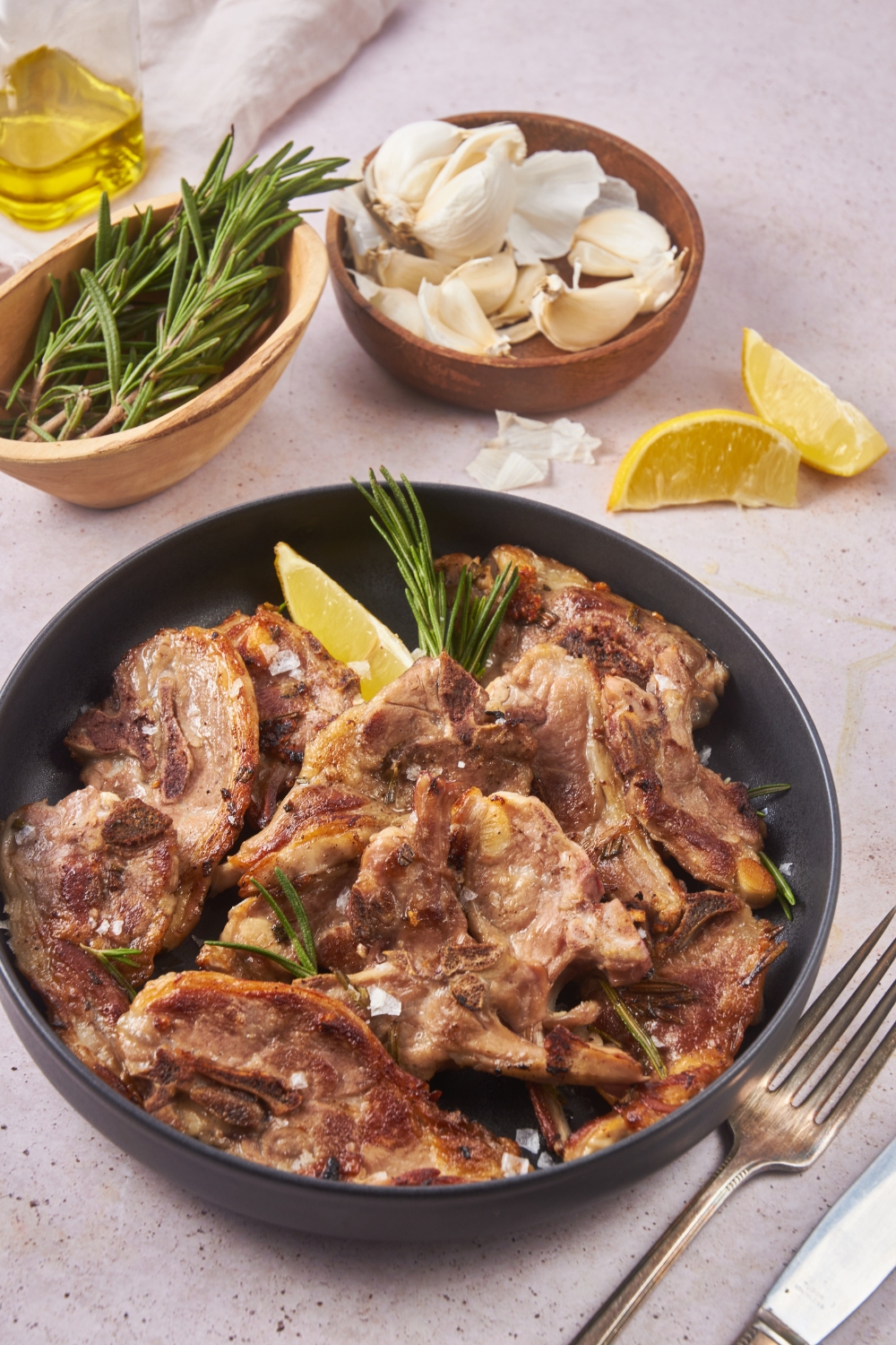A plate of cooked baby lamb chops piled on top of each other, garnished with sea salt and fresh herbs and a lemon wedge. Next to the plate is a fork, knife, and bowls of garlic and rosemary.
