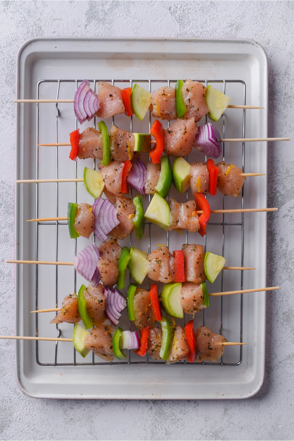 Six uncooked skewers of marinated chicken and vegetables on a wire rack atop a baking sheet.