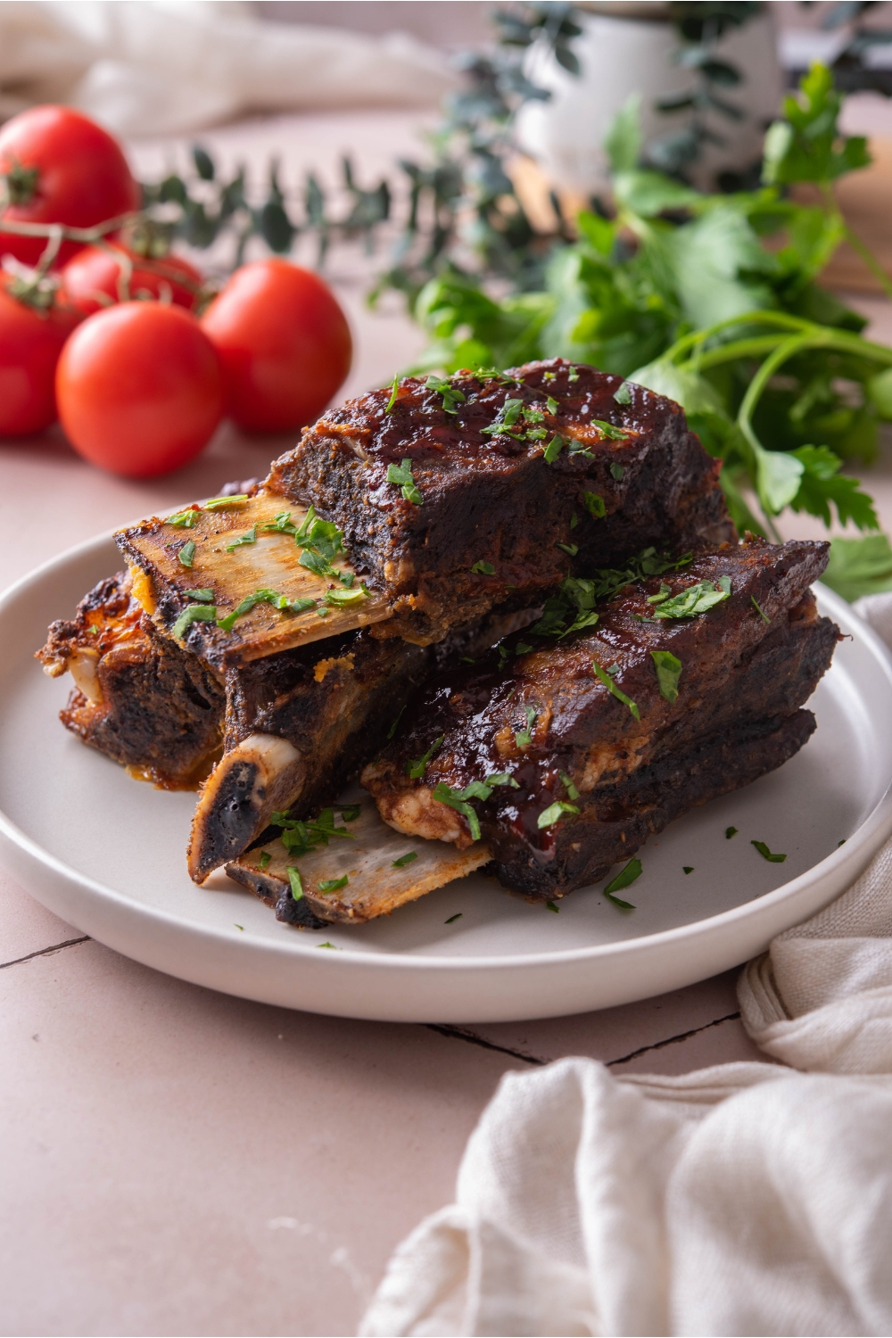 A stack of barbecue beef ribs on a white plate garnished with fresh herbs. In the background are vine tomatoes and green herbs.