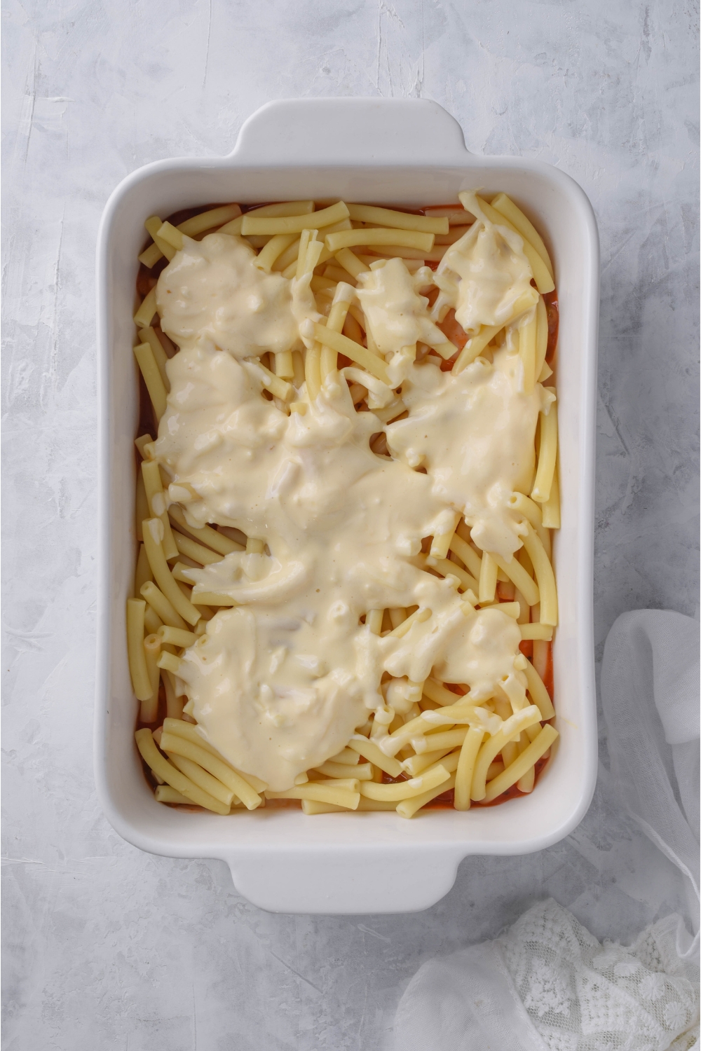 A white casserole dish filled with cooked ziti noodles and a creamy cheese sauce poured on top.