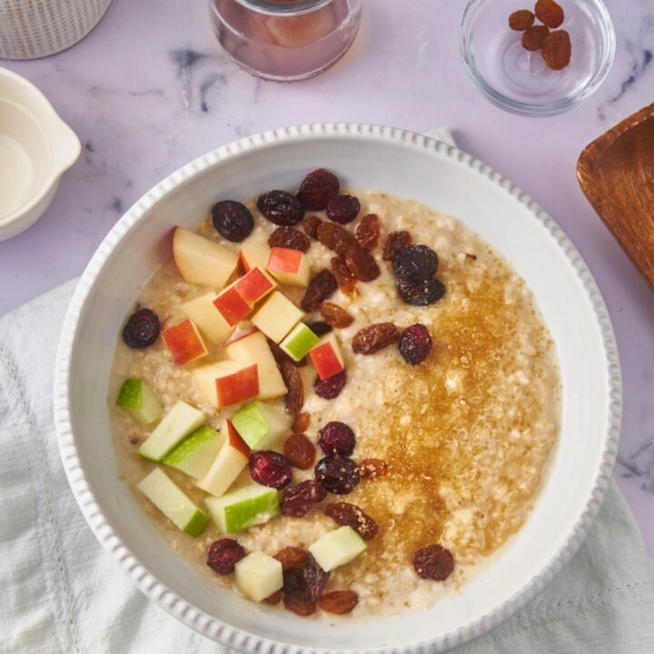 A bowl of oatmeal topped with brown sugar, raisins, and diced apples. Surrounding the oatmeal is a bowl of brown sugar, bowls of raisins, and a container of maple syrup.