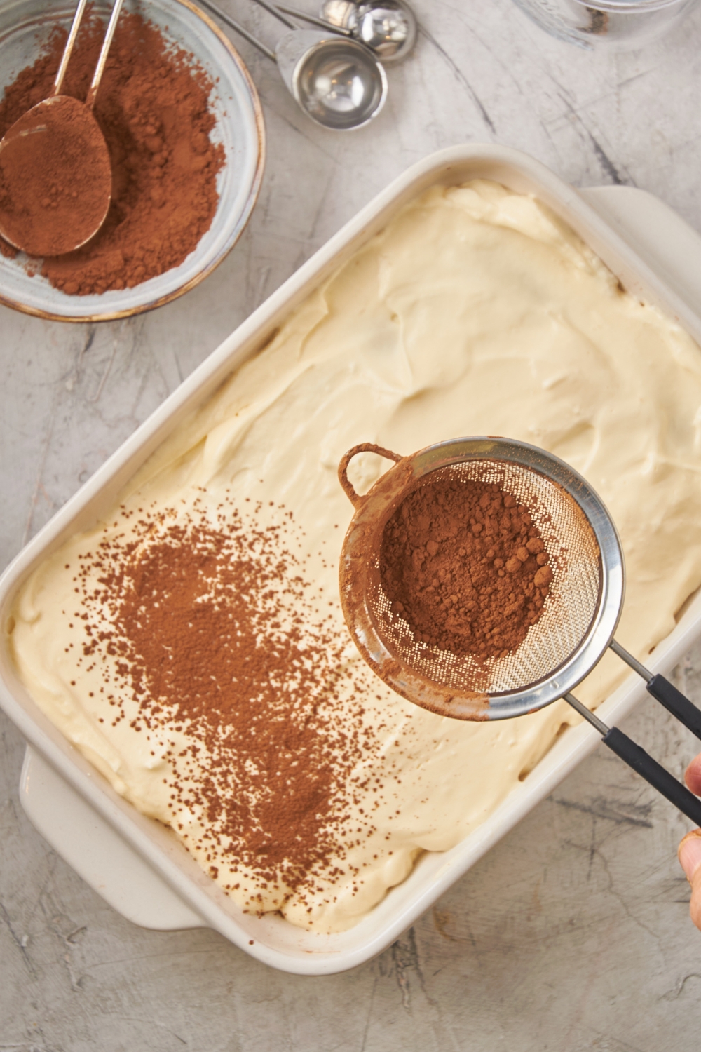 A pan with tiramisu, a sieve is sprinkling cocoa powder on top.