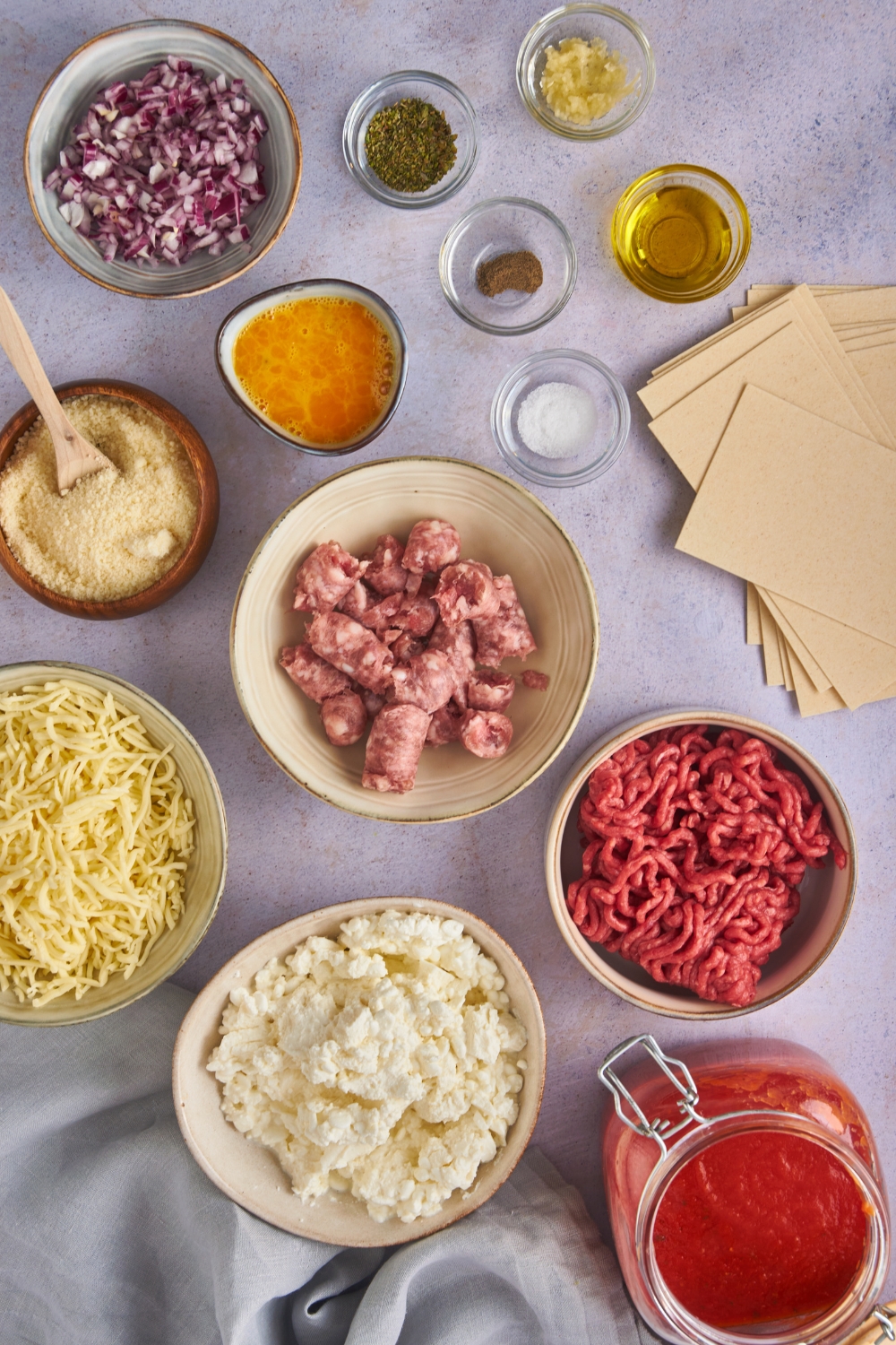 Overhead view of an assortment of ingredients including bowls of ground beef, sausage, diced red onion, garlic, a beaten egg, parmesan cheese, ricotta cheese, and tomato sauce.