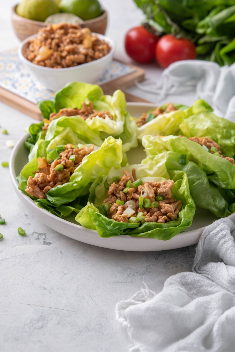 Five chicken lettuce wraps piled on a plate with a bowl of the chicken filling in the background.
