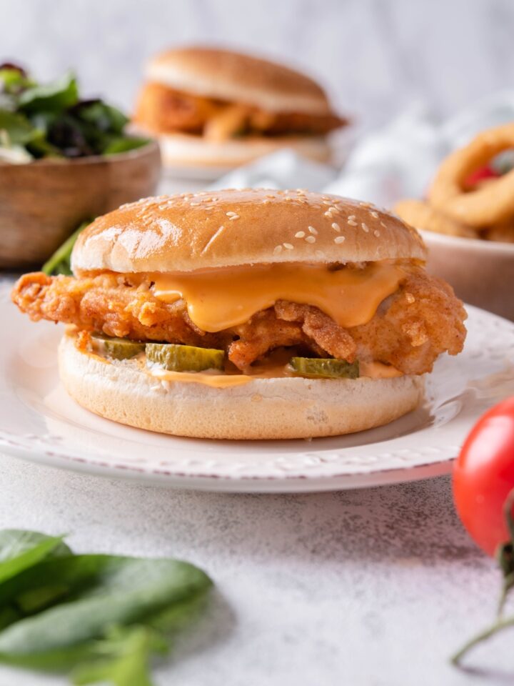 A fried chicken sandwich with spicy mayonnaise and pickles in a sesame seed bun. There is a second sandwich, a bowl of salad, and a bowl of onion rings in the background.