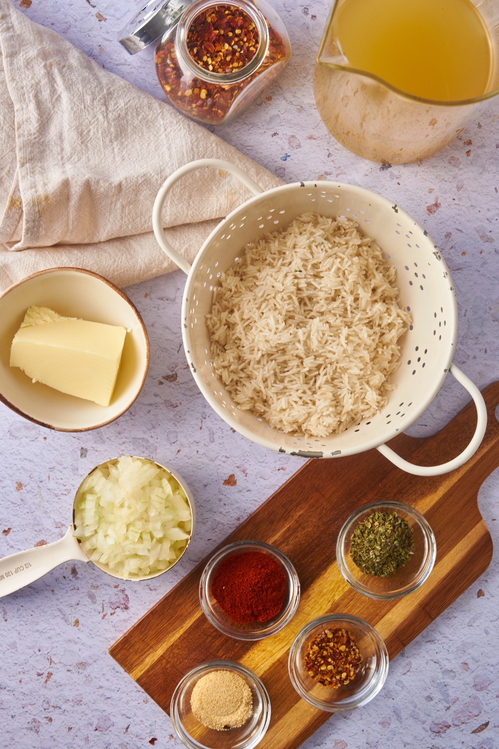 Overhead view of an assortment of ingredients including uncooked long grain rice in a colander and bowls of butter, diced onion, spices, and a pitcher of chicken stock.