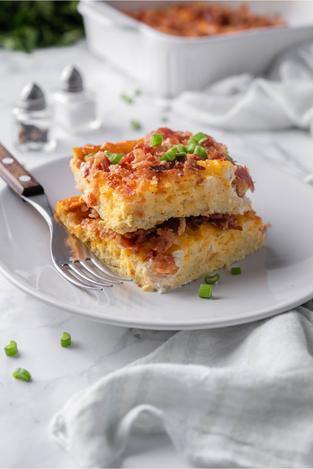Two squares of egg and bacon breakfast casserole stacked on top of each other and garnished with green onions. There is a fork on the plate.