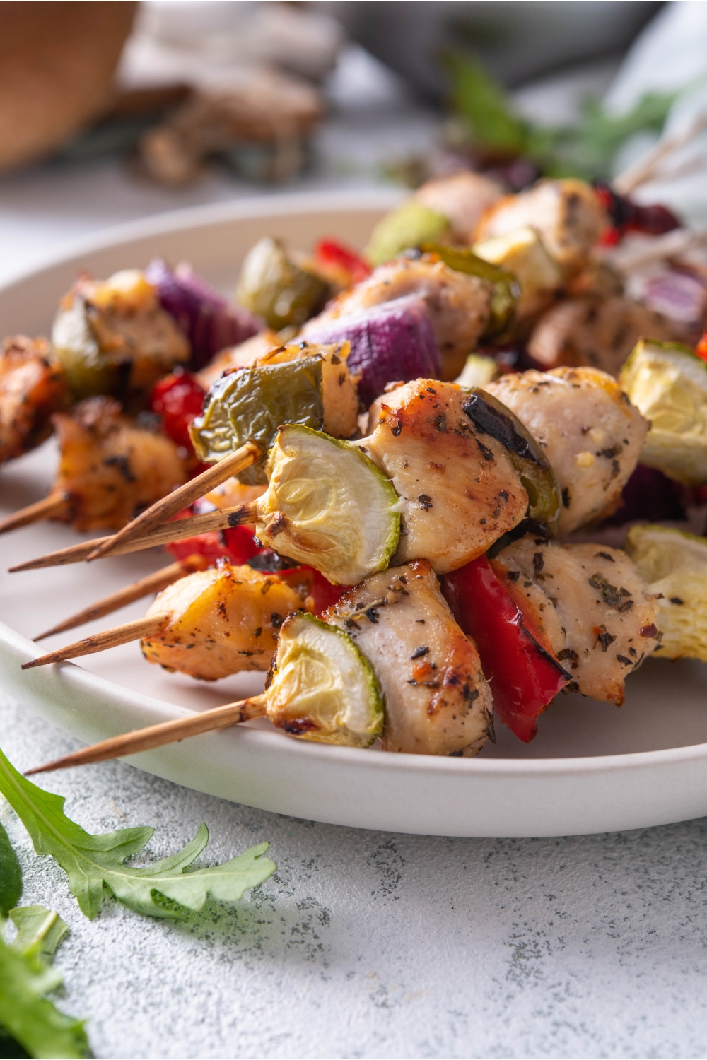Close up of a plate of six chicken and vegetable skewers with red onion, bell pepper, and zucchini in the skewers.
