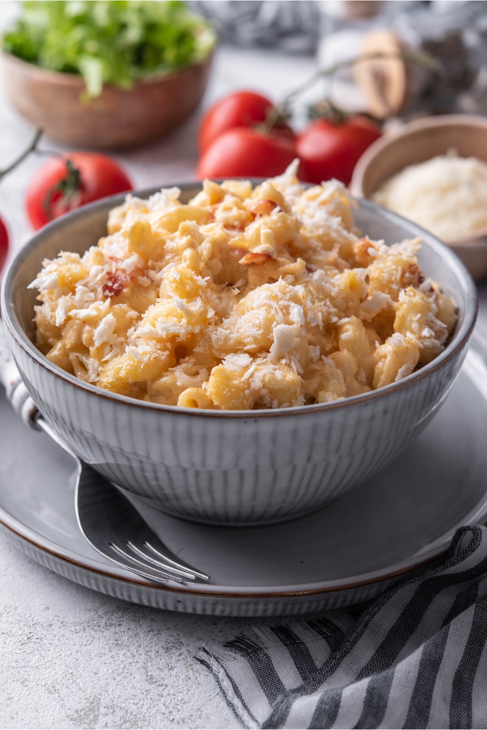 A bowl of mac and cheese with crumbled bacon and bread crumbs on top. The mac and cheese is in a blue bowl atop a blue plate with a fork on it.