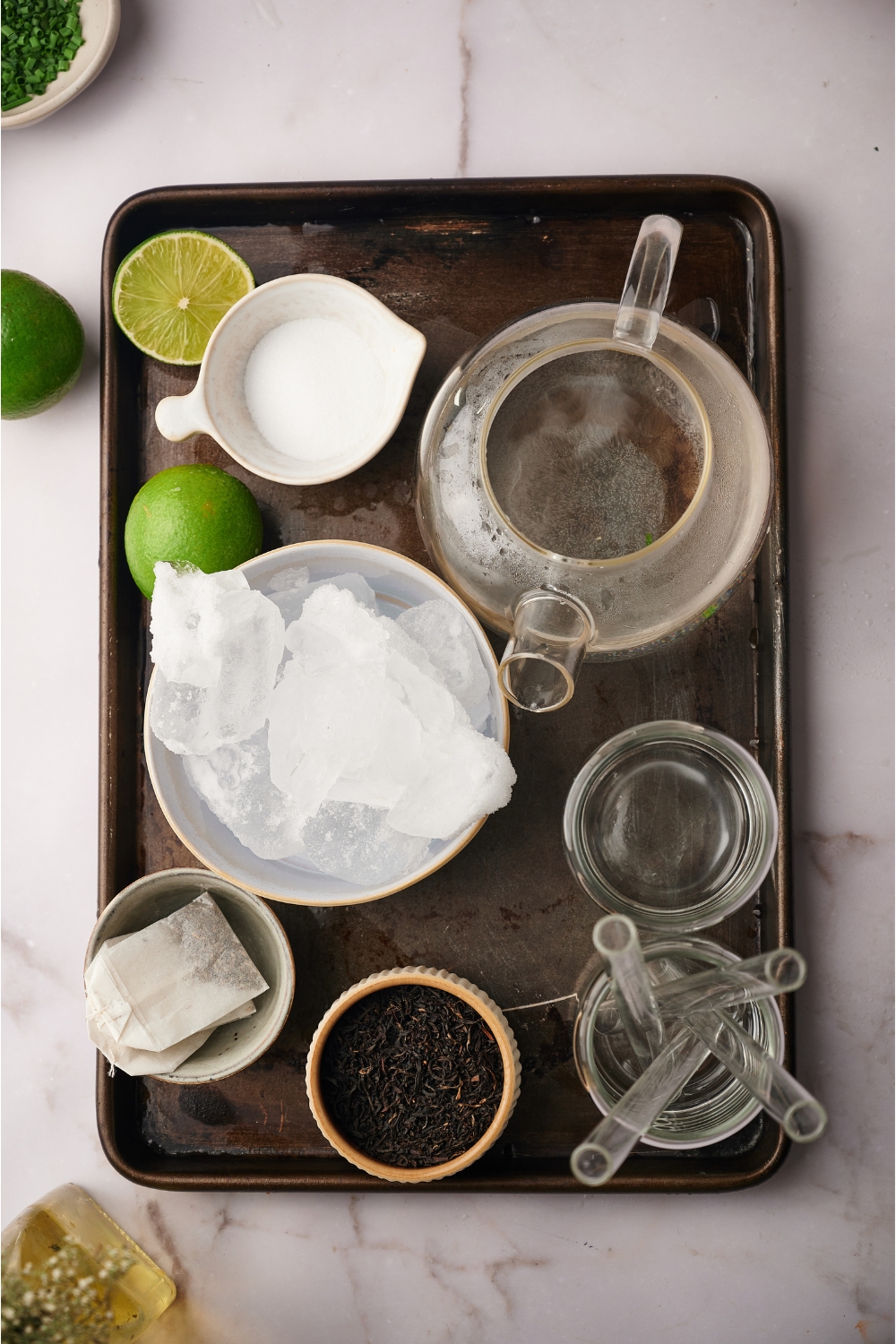 Overhead view of a tray with a tea pot, a bowl of ice, a bowl of sugar, tea bags, limes, an empty glass, and a glass filled with straws.