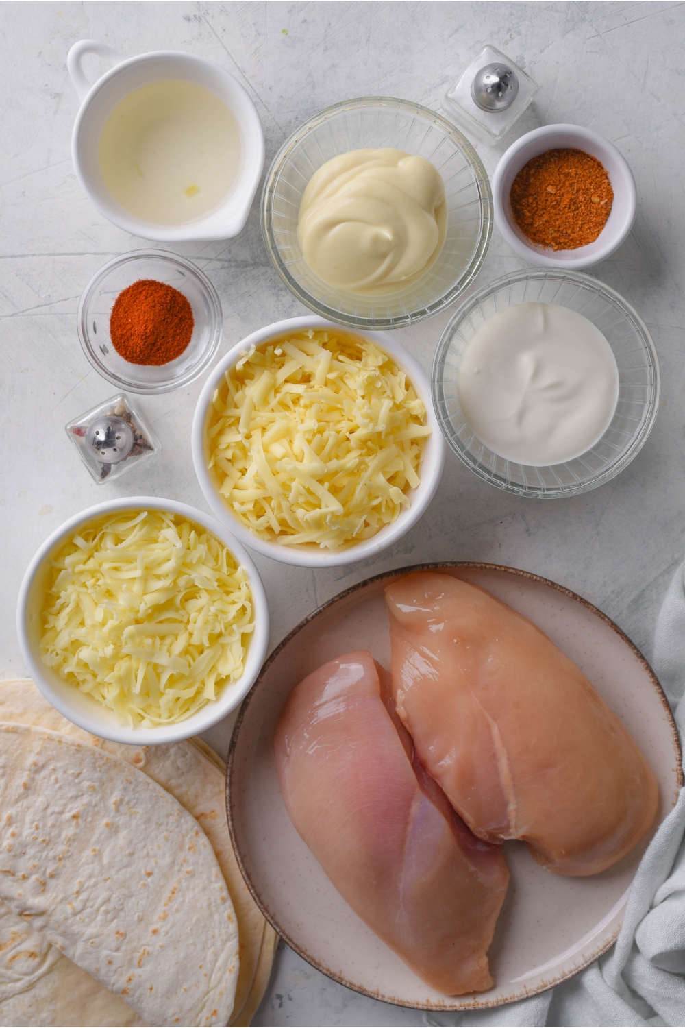 An assortment of ingredients including a plate of chicken breasts and bowls of mayonnaise, shredded cheese, spices, sour cream, and a pile of tortillas.