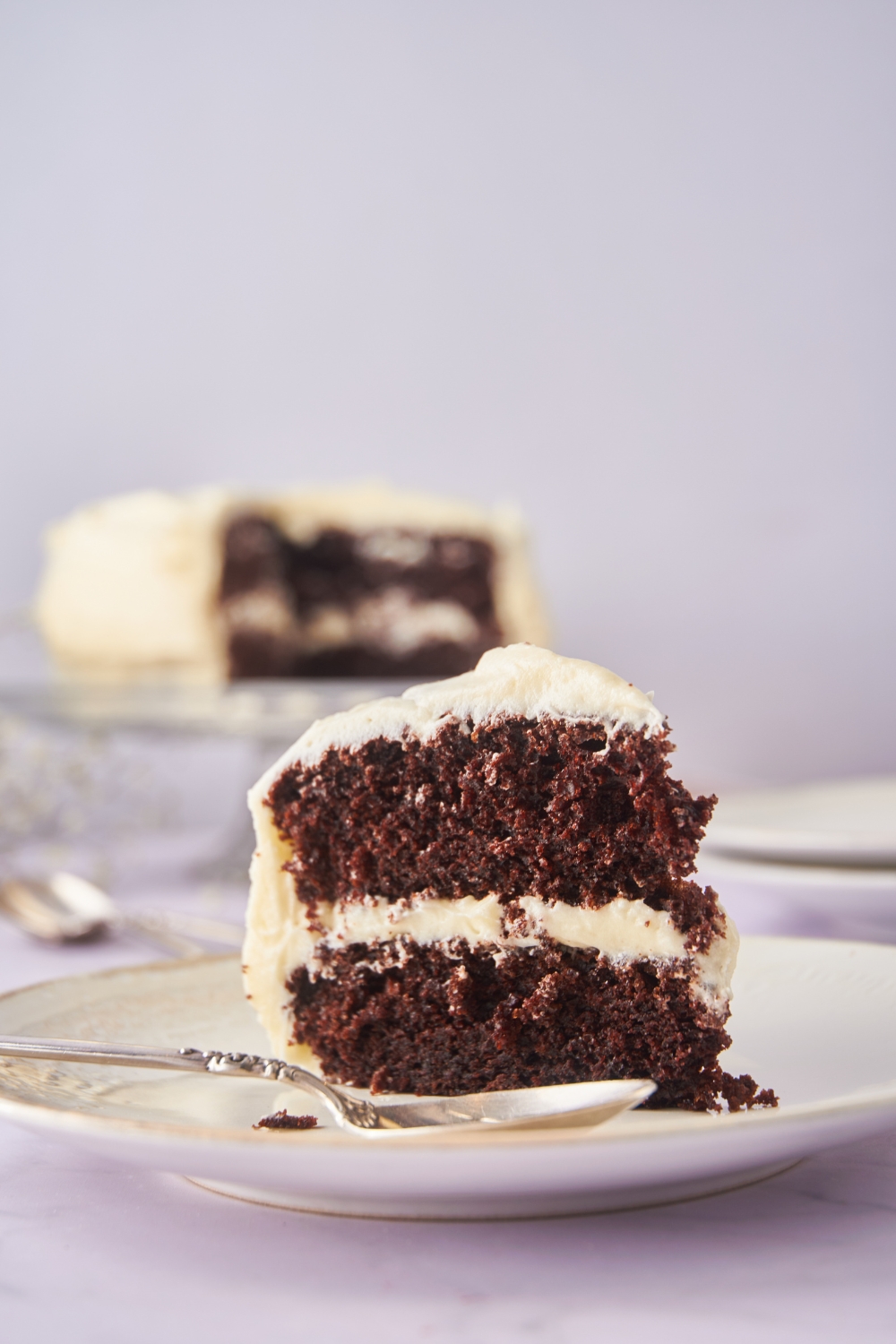 A slice of chocolate cake layered with cream cheese frosting on a plate with a spoon next to it.