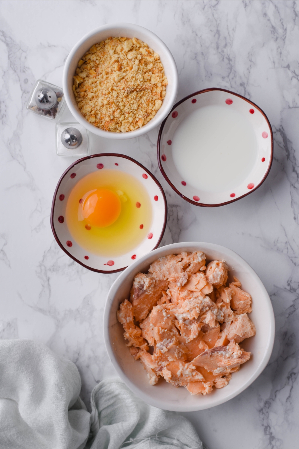 Overhead view of an assortment of ingredients including bowls of canned salmon, an egg, milk, cracker crumbs, and mini salt and pepper shakers.