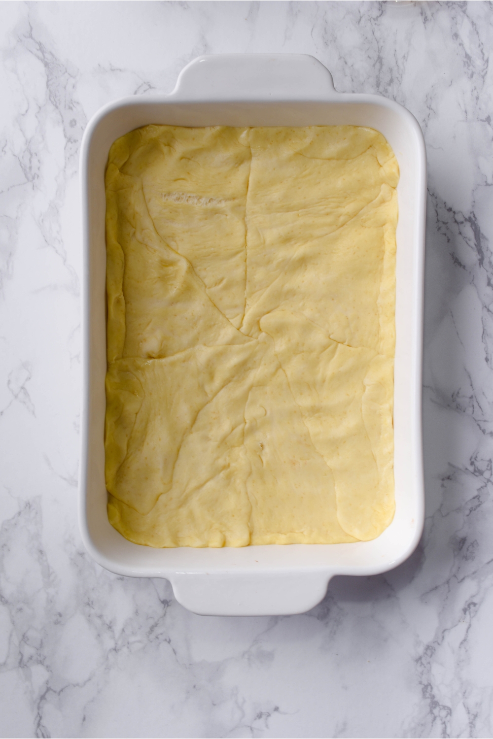 A white baking dish with the bottom covered by a flat sheet of crescent roll dough.