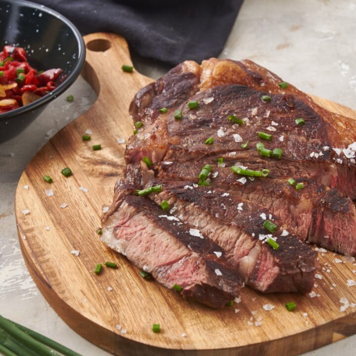 A ribeye steak sliced thin on a wooden board with flaky sea salt and chopped chives. Surrounding the board is a bowl of garlic, a bunch of chives, and a bottle of oil.