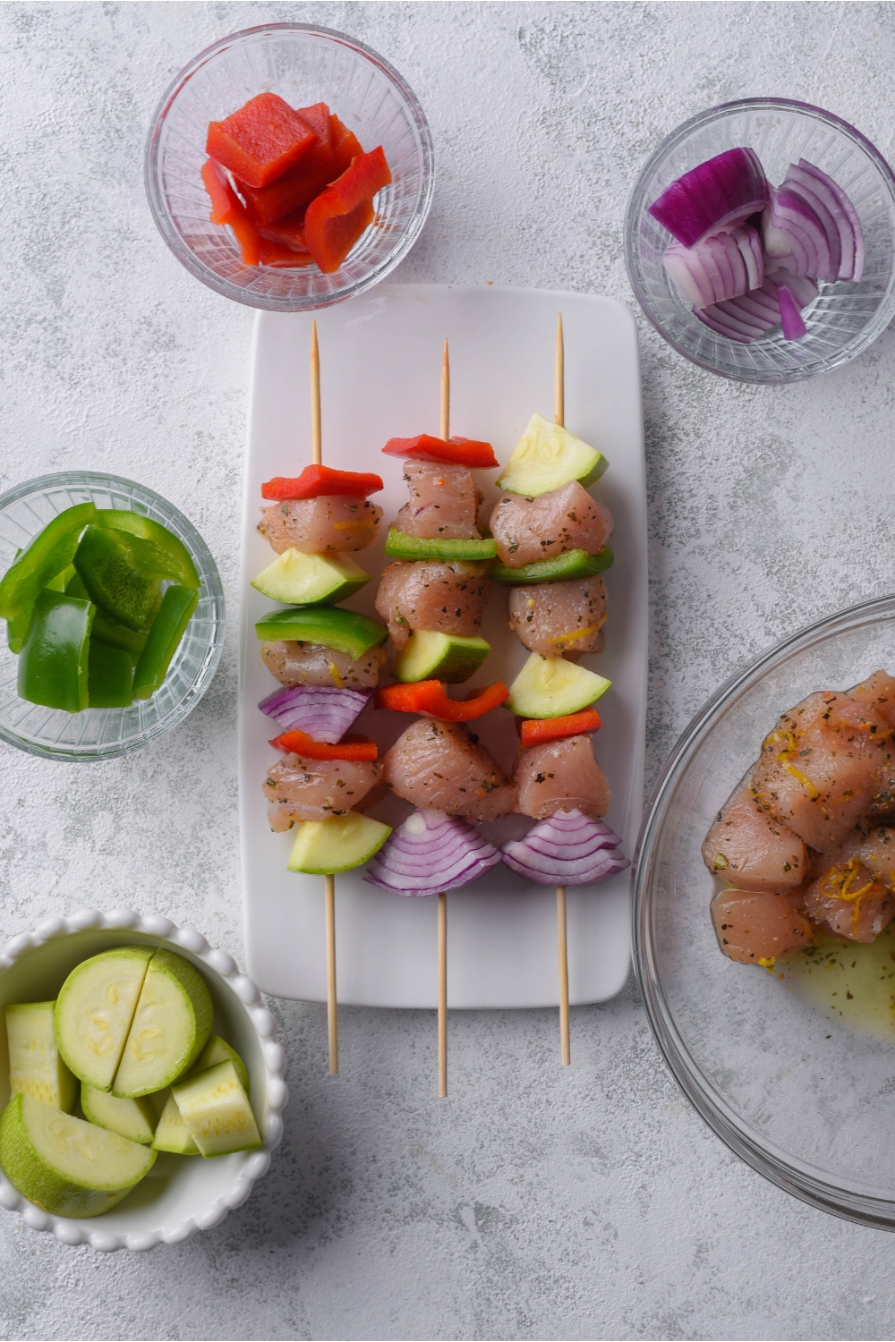 Three skewers of alternating pieces of raw marinated chicken, red onion, zucchini, and red bell peppers on a white plate, surrounded by bowls of the skewer ingredients.