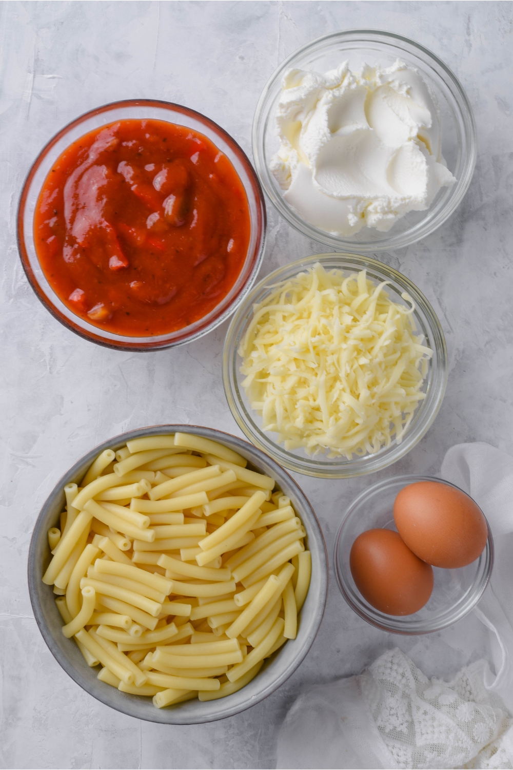 An assortment of ingredients including bowls of ricotta cheese, mozzarella cheese, tomato sauce, cooked ziti pasta, and two eggs.