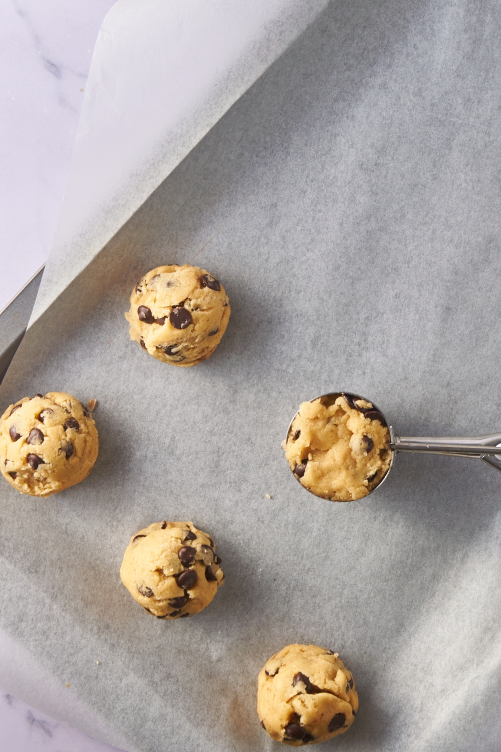 Cookie dough balls being portioned with an ice cream scoop onto a baking sheet lined with parchment paper.