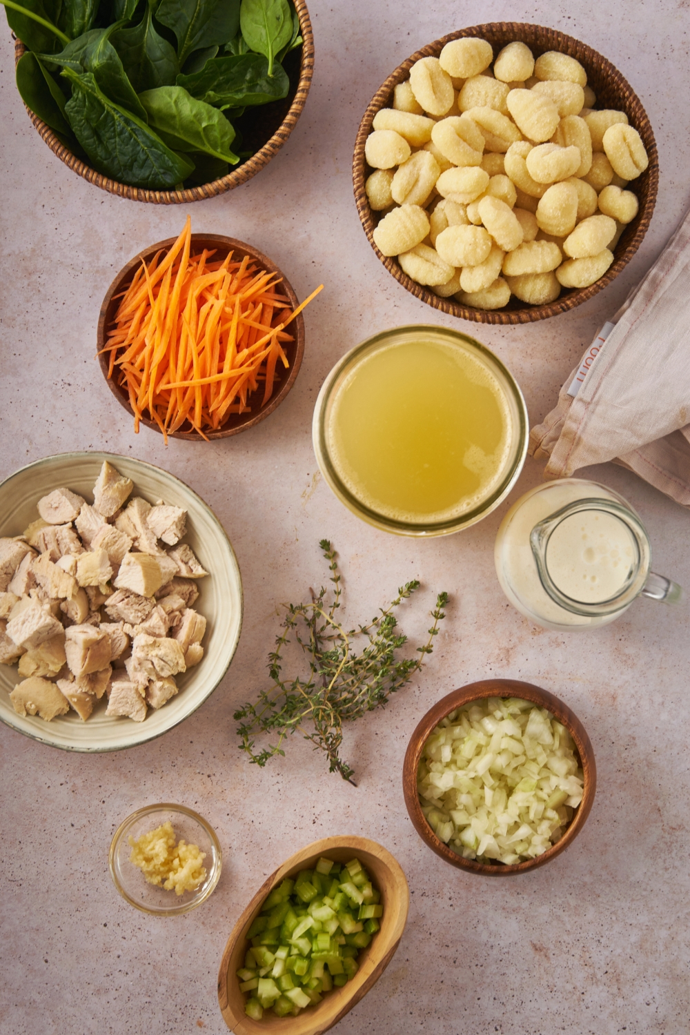 Overhead view of an assortment of ingredients including bowls of shredded carrots, potato gnocchi, diced chicken, diced onion, diced celery, and chicken broth.