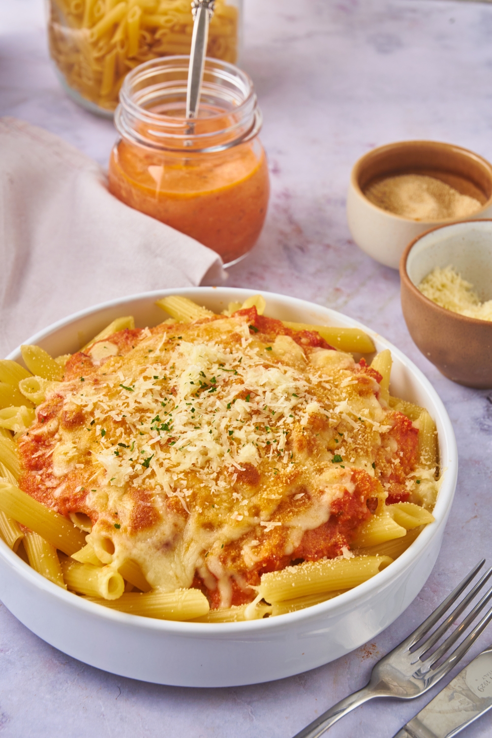 A bowl of ziti topped with a creamy marinara sauce, shredded cheese, and fresh herbs. There is a bowl of creamy marinara and small bowls of shredded cheese next to it.