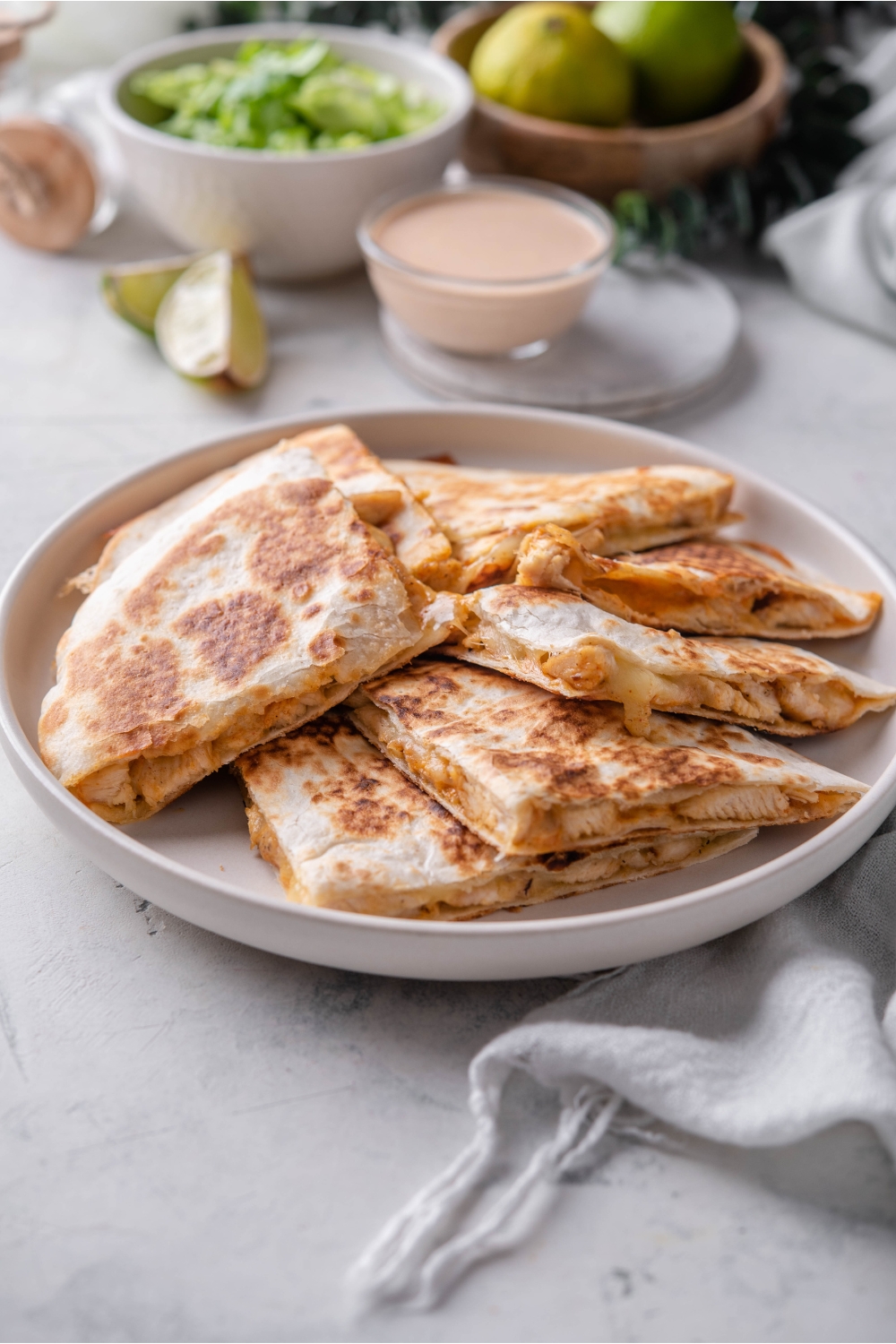 A plate of chicken quesadilla wedges piled on top of each other with a bowl of quesadilla sauce and a bowl of lettuce in the background.
