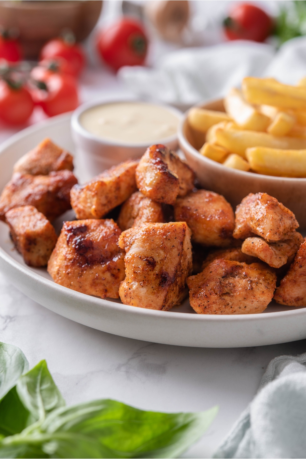 A pile of grilled chicken nuggets on a white plate with a side of french fries and a bowl of dipping sauce.