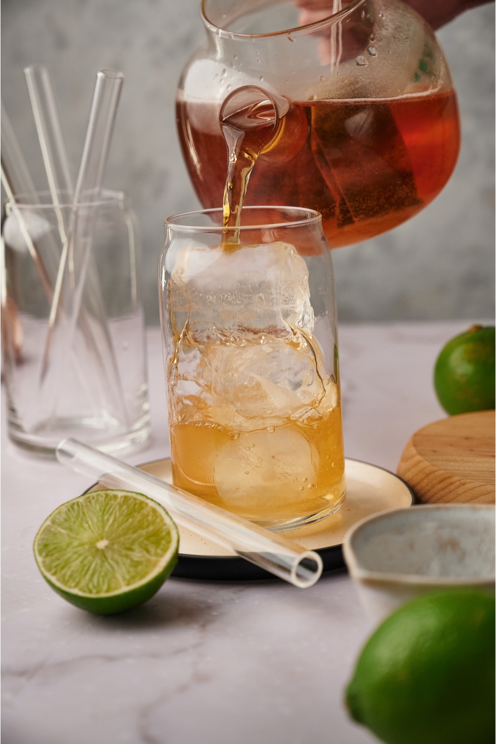 A glass filled with ice and a teapot pouring tea into the glass. There is a straw and a halved lime next to the glass.