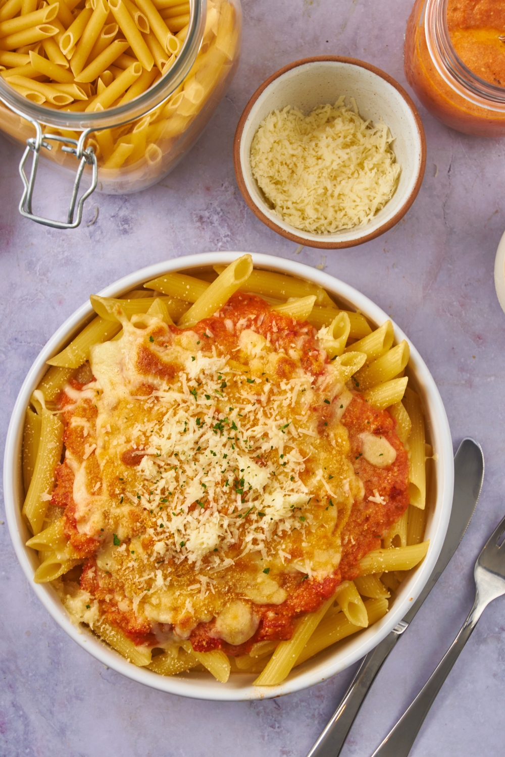 Overhead view of a bowl of ziti topped with a creamy marinara sauce, shredded cheese, and fresh herbs. There is a bowl of shredded cheese next to the pasta.
