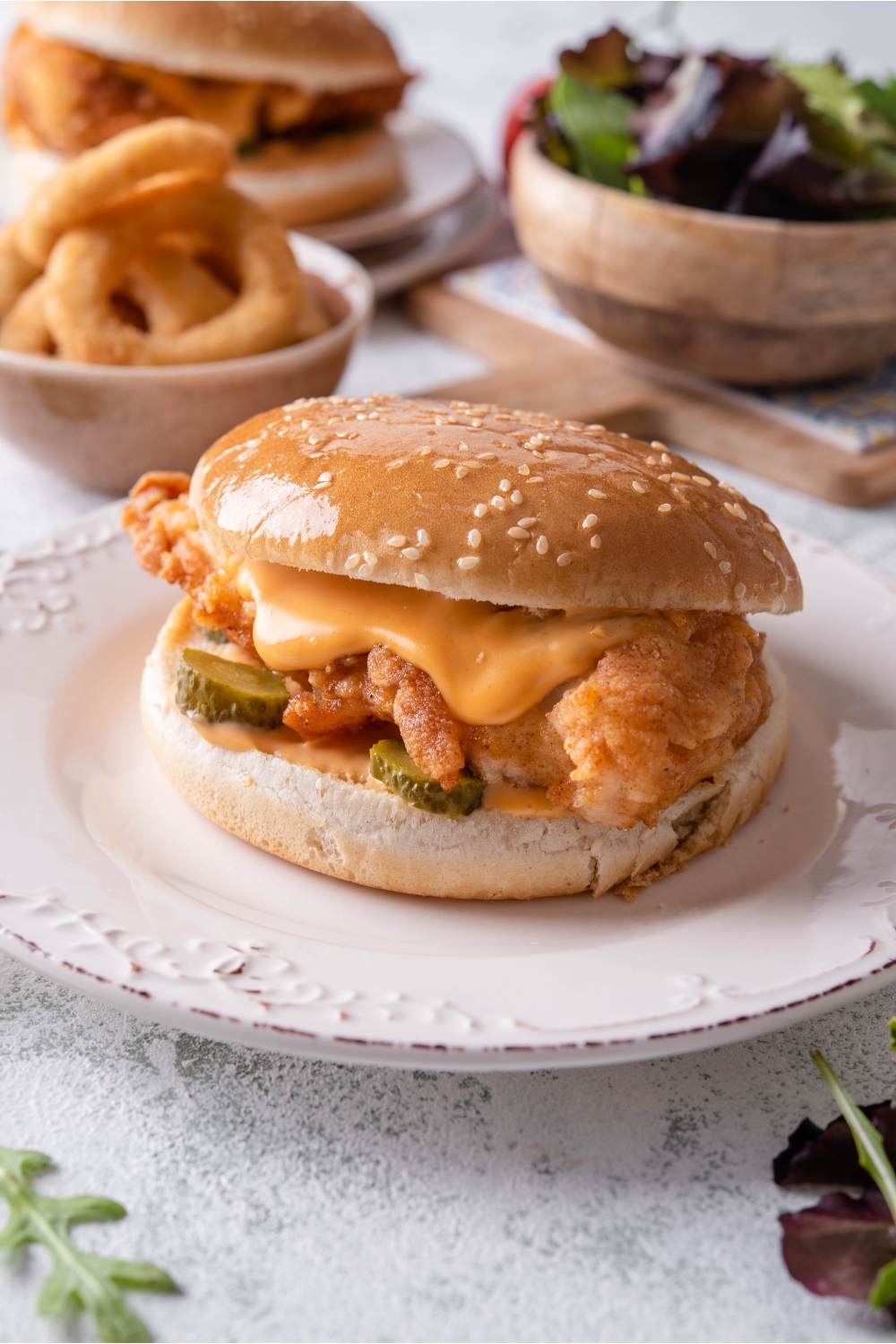 A fried chicken sandwich with spicy mayonnaise and pickles in a sesame seed bun. There is a second sandwich, a bowl of salad, and a bowl of onion rings in the background.