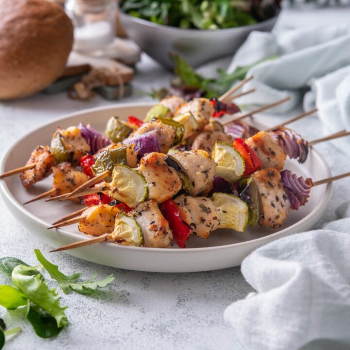 A plate of six chicken and vegetable skewers with red onion, bell pepper, and zucchini in the skewers.
