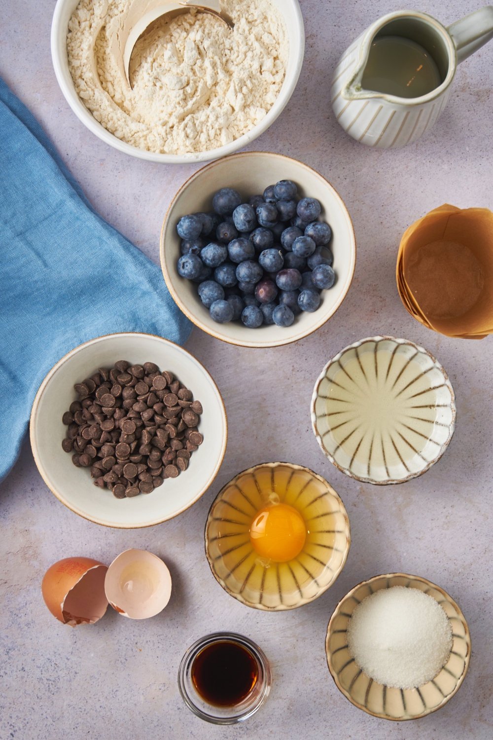 Overhead view of an assortment of ingredients including bowls of blueberries, chocolate chips, an egg, sugar, oil, baking mix, and vanilla extract.