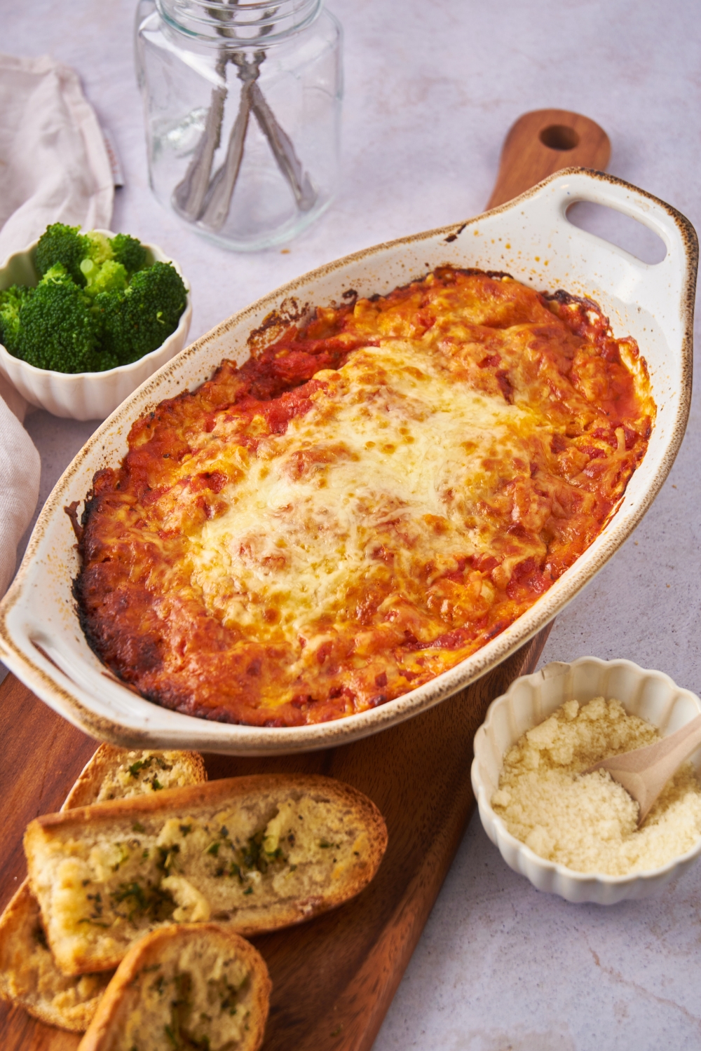 Baking dish filled with baked ravioli covered in melted cheese on a wooden board with garlic bread and a bowl of parmesan cheese.