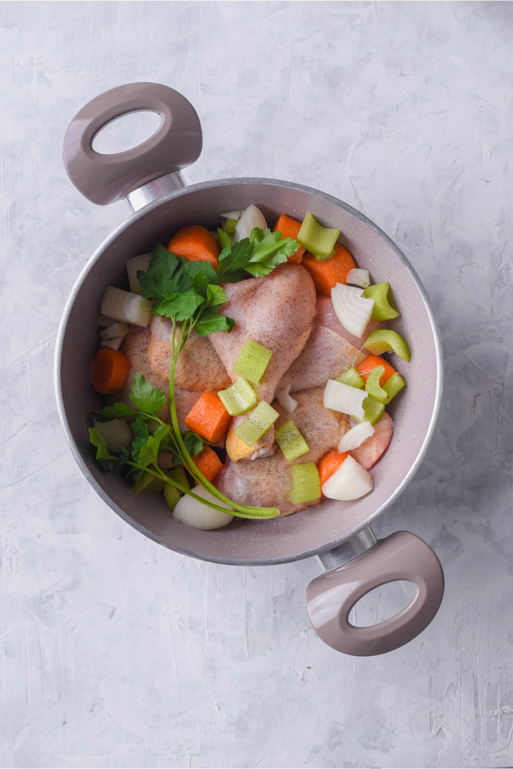 Grey pot filled with raw seasoned chicken topped with fresh celery, carrots, onions, and parsley.